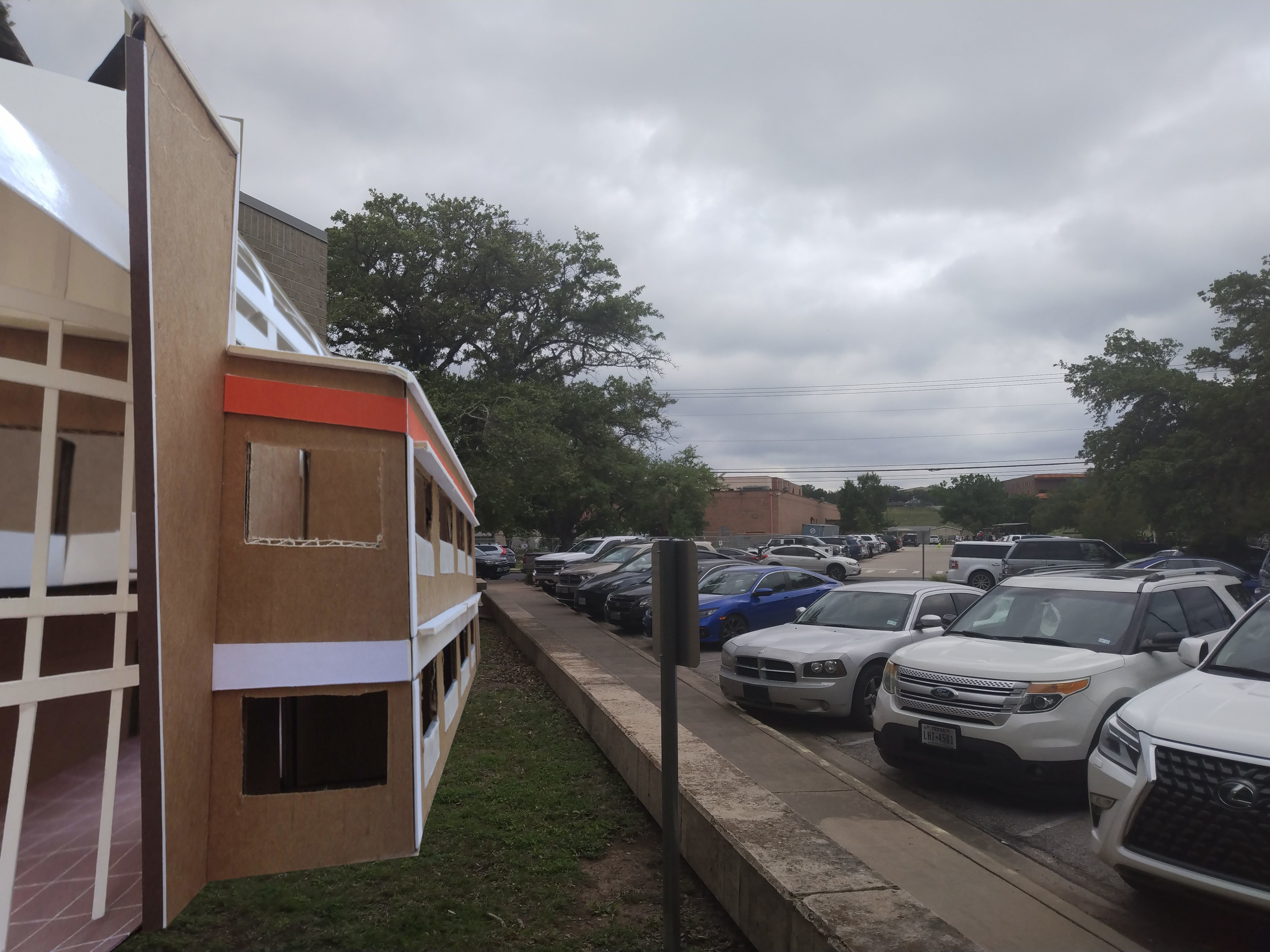 a photoshopped image of the cardboard model standing to scale on the left of a real parking lot