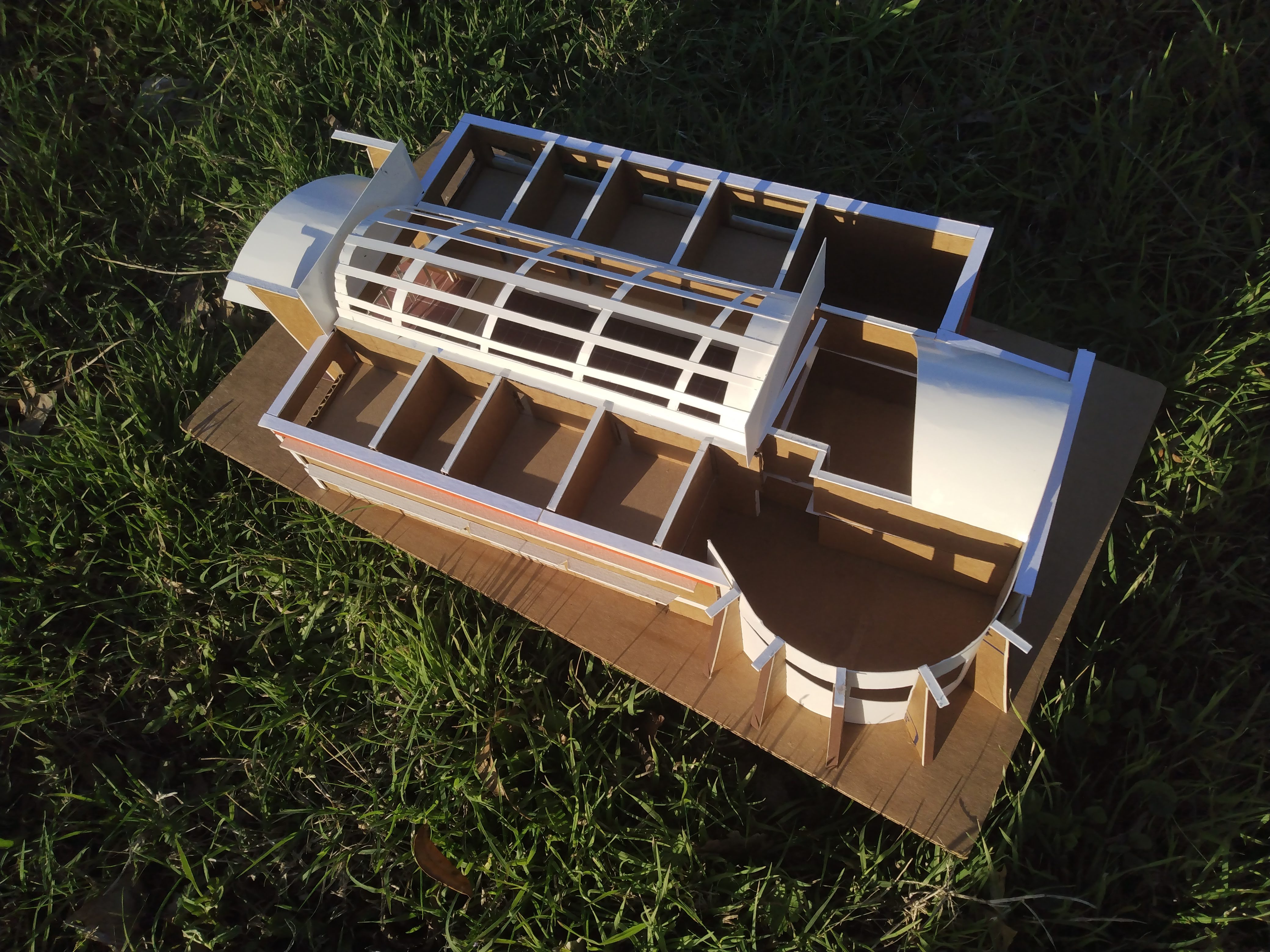 the cardboard mockup is on a grassy field with the arc-shaped roof and second-story classrooms visible from above